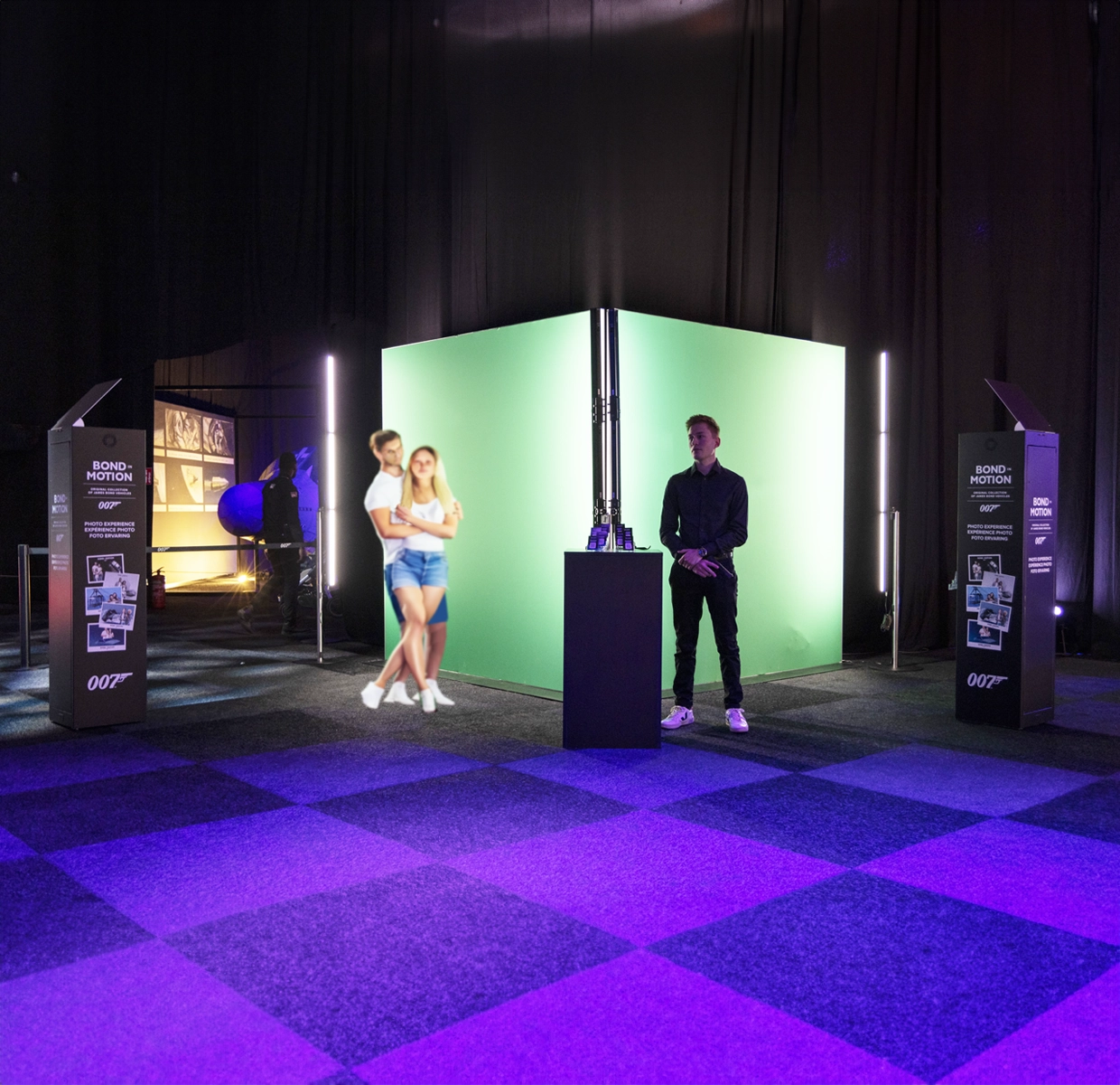 A couple posing for a photo in front of a green screen at the 'Bond in Motion' exhibit's automated photo studio