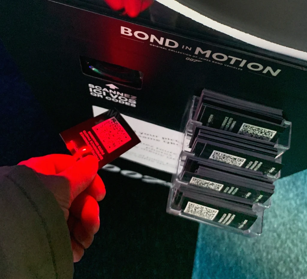 A visitor's hand holding a QR code card at the 'Bond in Motion' exhibit, ready to interact with the scan and capture system
