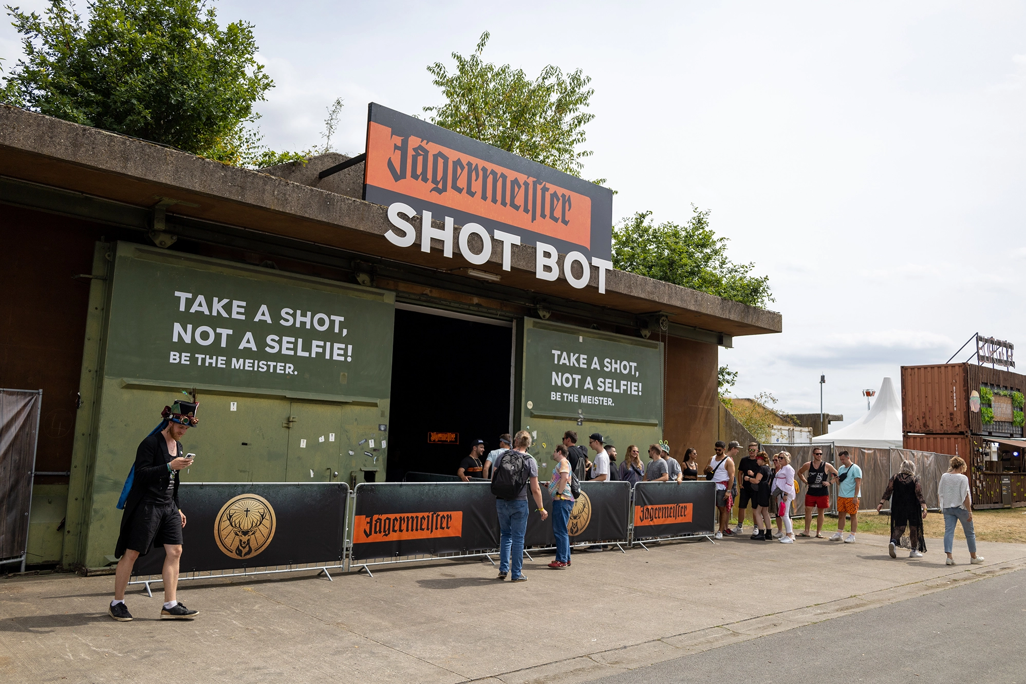 Attendees line up at a unique outdoor activation with the bold slogan 'TAKE A SHOT, NOT A SELFIE!' prominently displayed, featuring interactive engagement with a lively atmosphere.