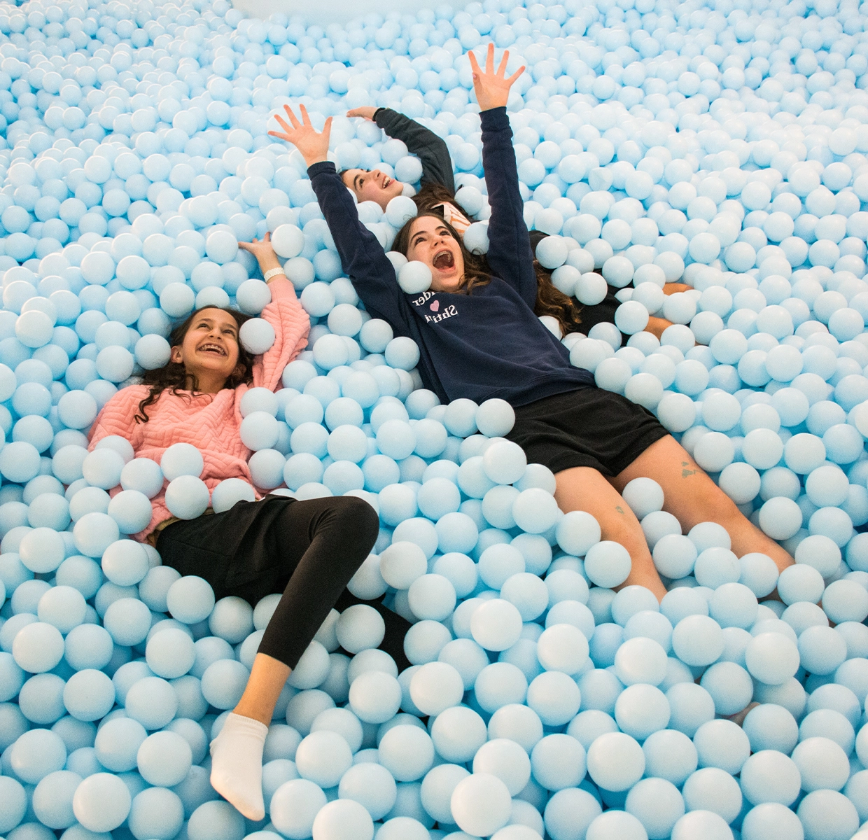 Two girls lie back in a sea of pale blue balls, laughing and creating memories at the Color Factory's interactive ball pit exhibit