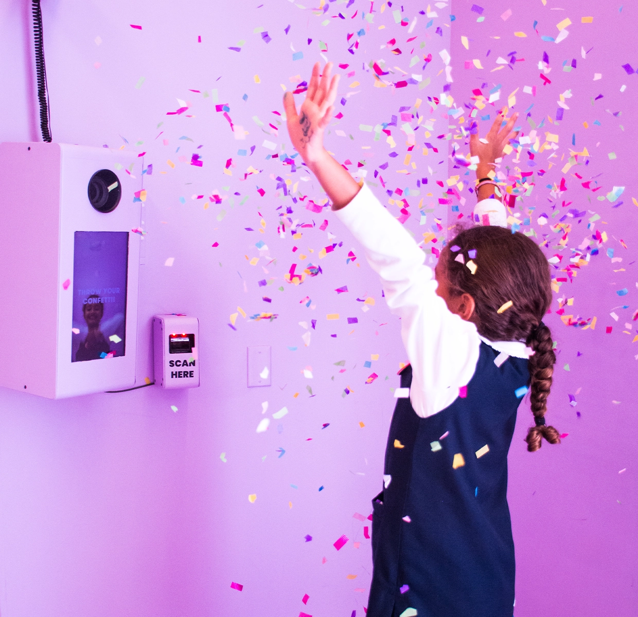 A girl throws confetti in the air, capturing a magical moment against a purple backdrop with a QR code for photo retrieval at the Color Factory