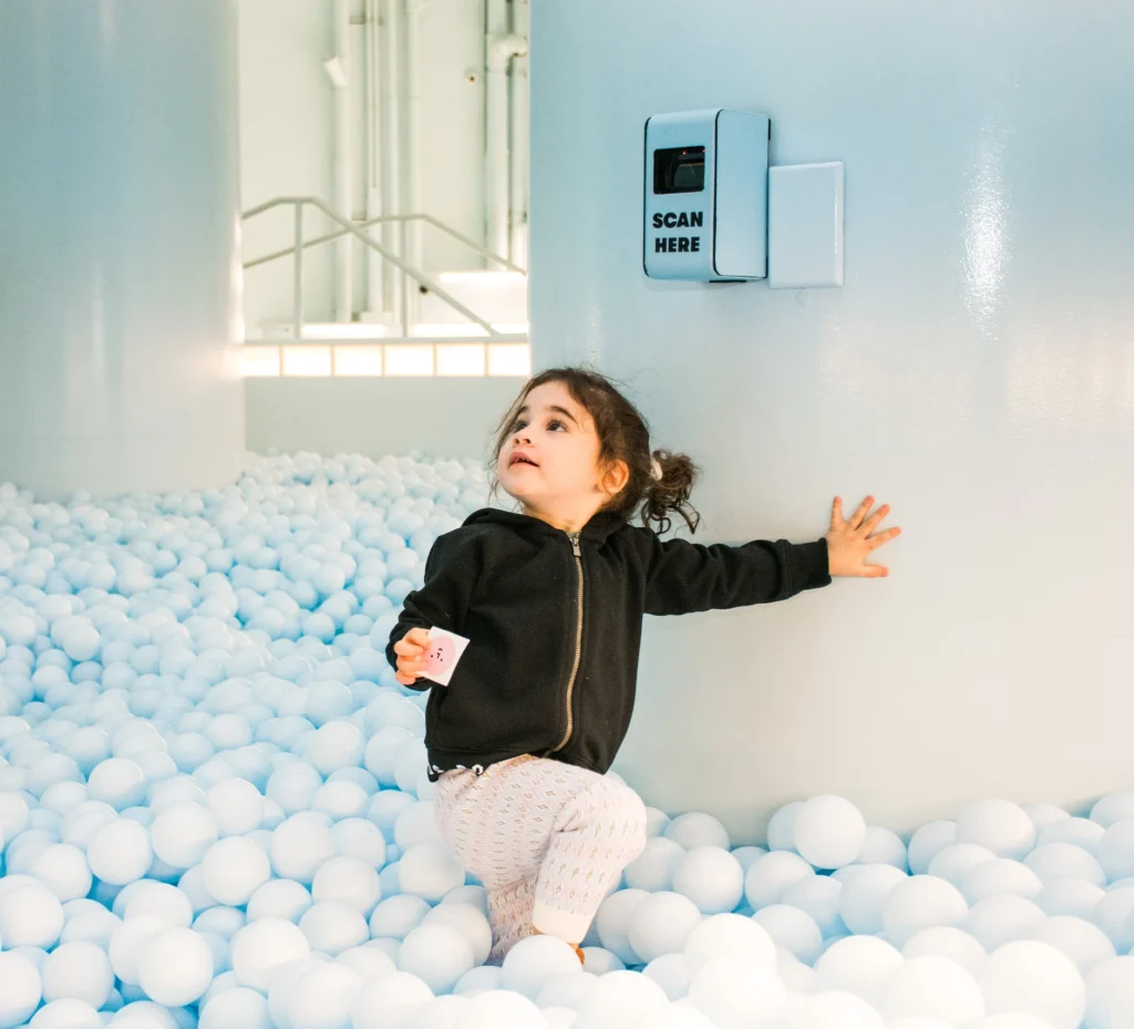 A young child enjoys the sensory delight of a pale blue ball pit, with easily accessible QR codes for capturing the joyous moment at the Color Factory